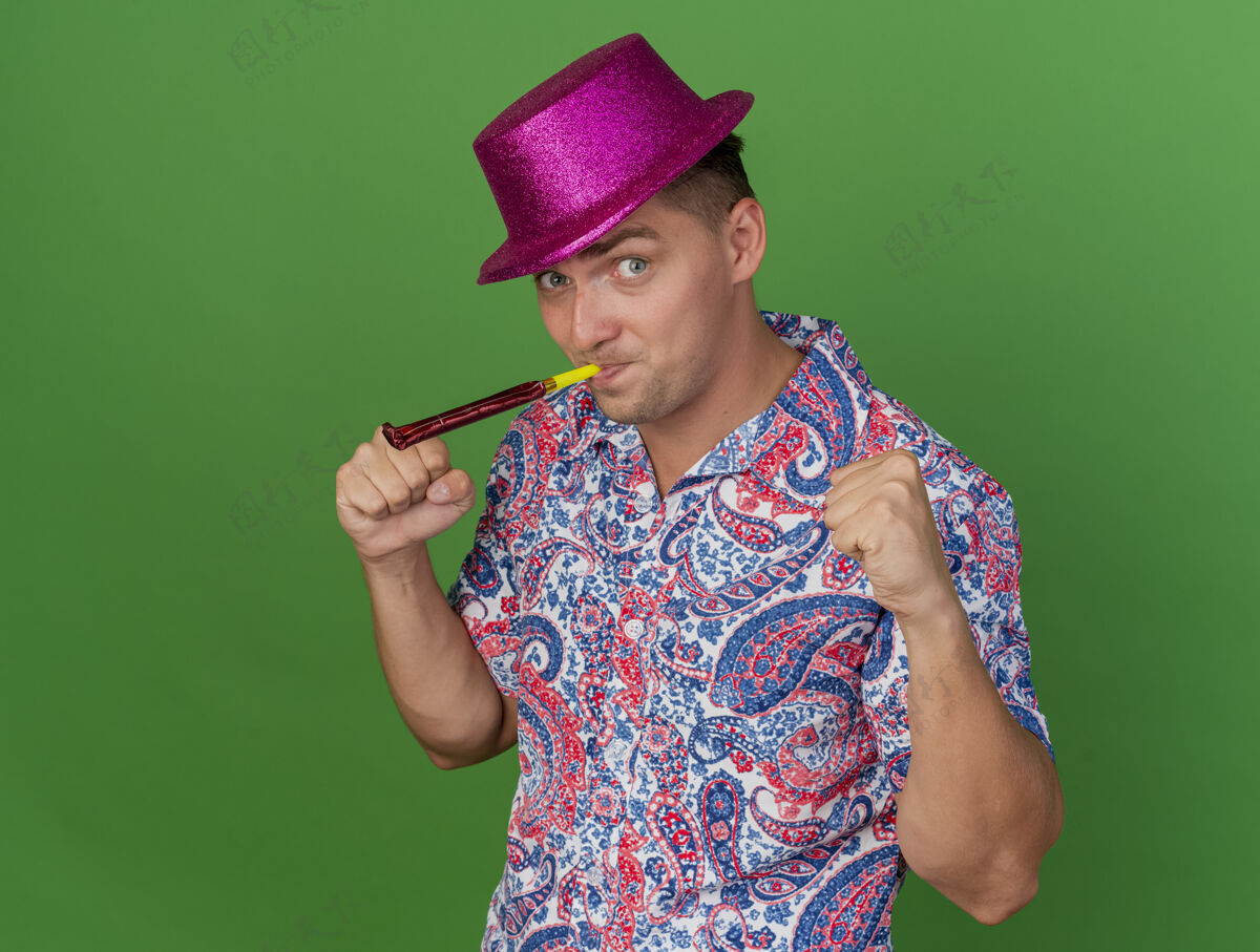 穿快乐的年轻人戴着粉红色的帽子吹派对吹风机 在绿色背景上显示“是”的手势帽子欢乐吹