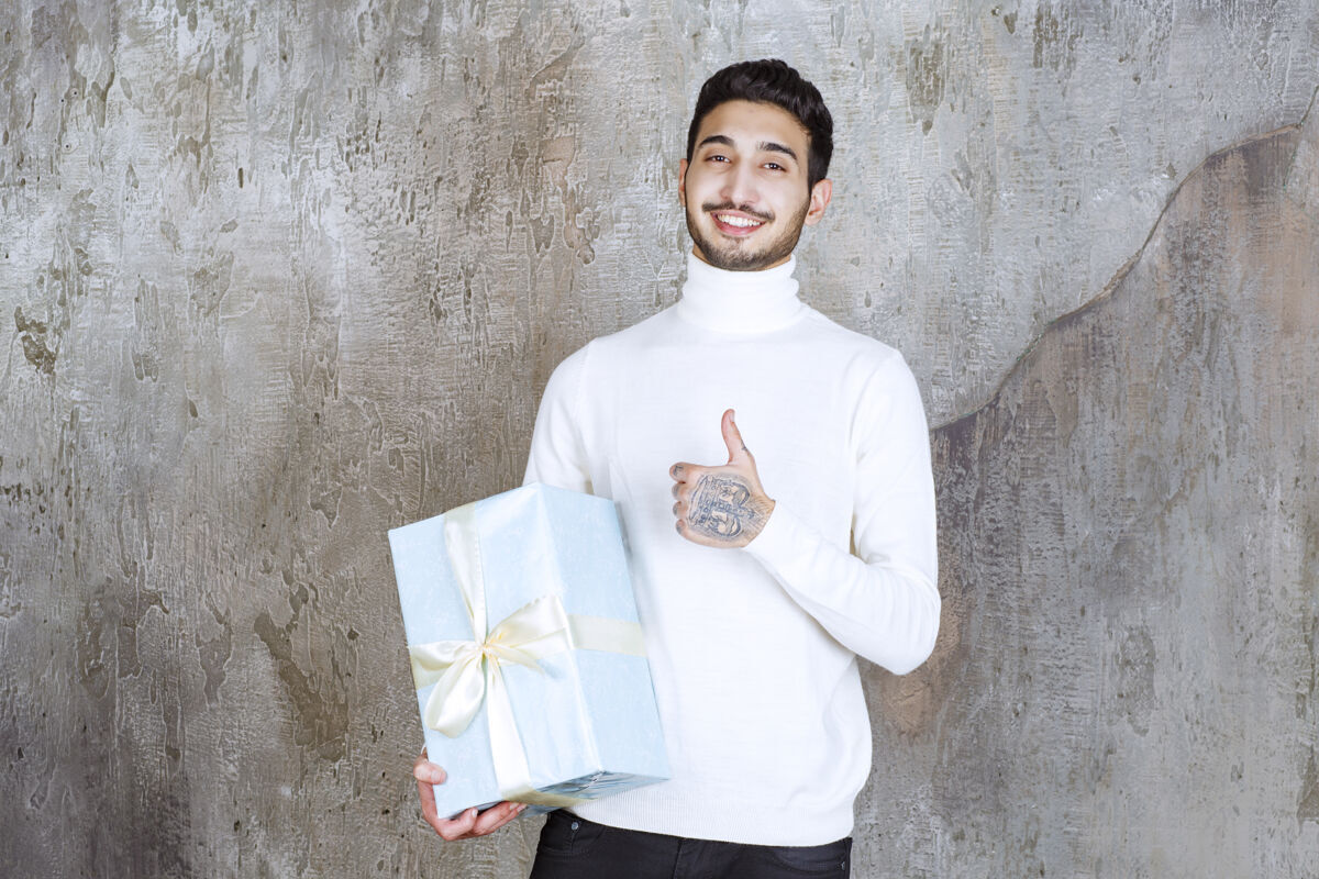 人一个穿着白色毛衣的男人手里拿着一个蓝色的礼盒 并显示出积极的手势礼物年轻成就