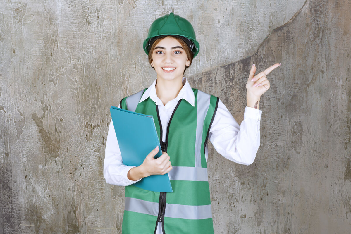 人身着绿色制服 头戴安全帽的女工程师手持绿色项目文件夹 指着右侧工作人员方向检查表