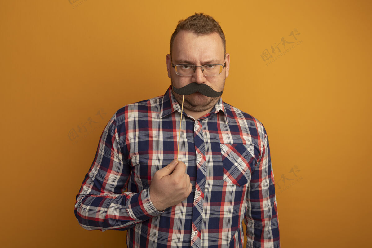 胡子一个戴着眼镜和格子衬衫的男人站在橙色的墙上 手里拿着一把滑稽的小胡子拿着搞笑眼镜