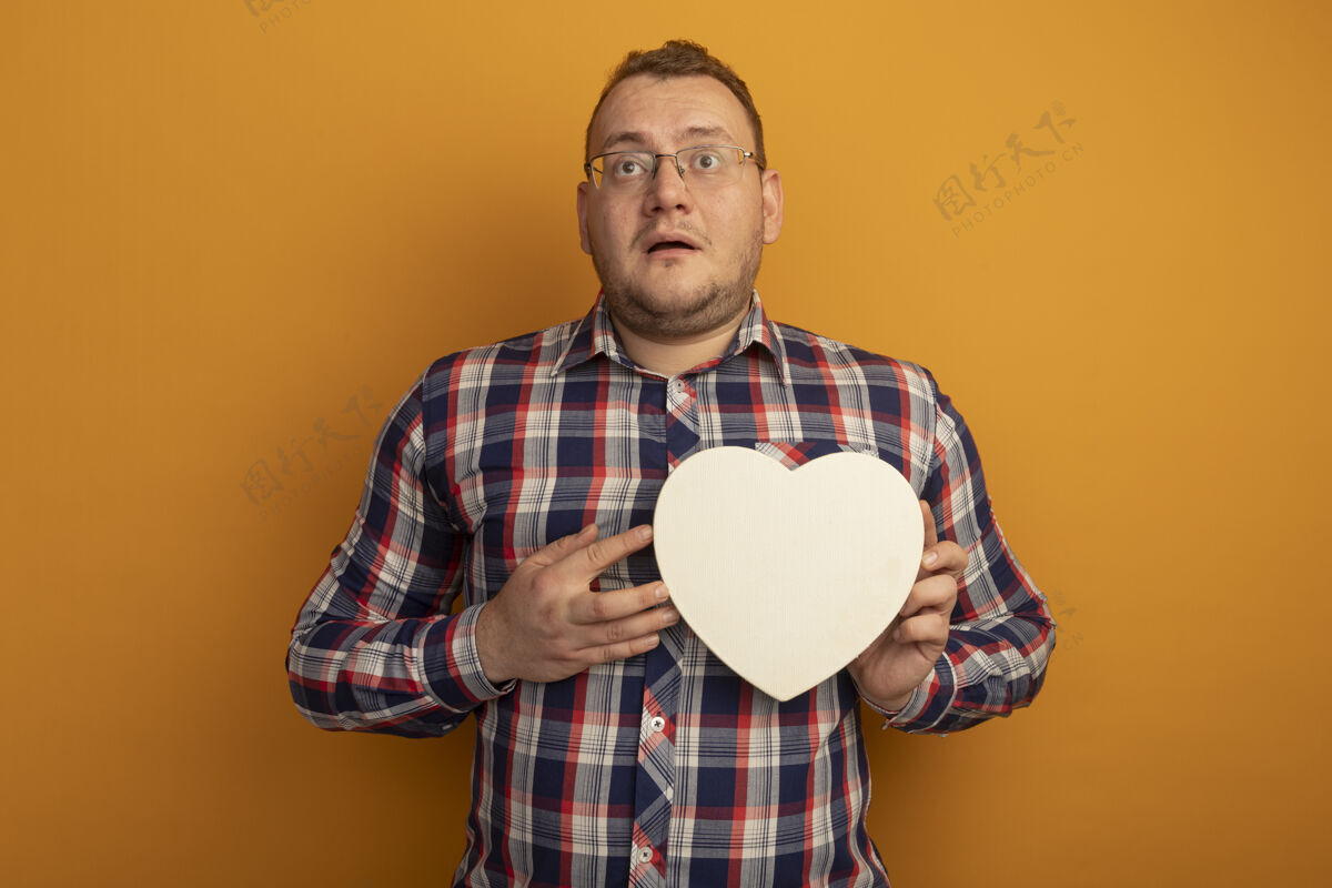 检查一个戴着眼镜和格子衬衫的男人抱着cardborad的心站在橙色的墙上困惑地看着一边站着放一边心