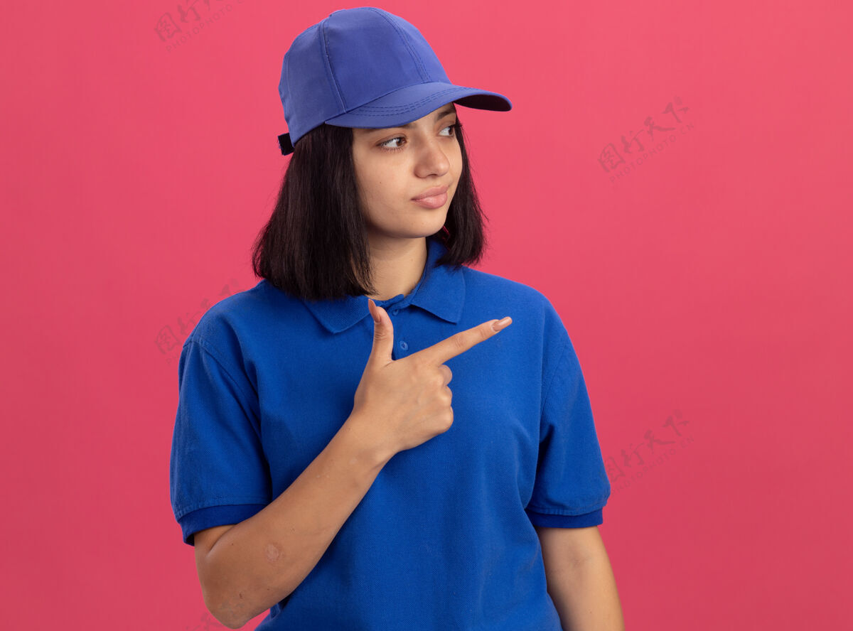 壁板身穿蓝色制服 头戴鸭舌帽的年轻送货员站在粉红色的墙上 自信地指着旁边的指示牌目录女孩指向