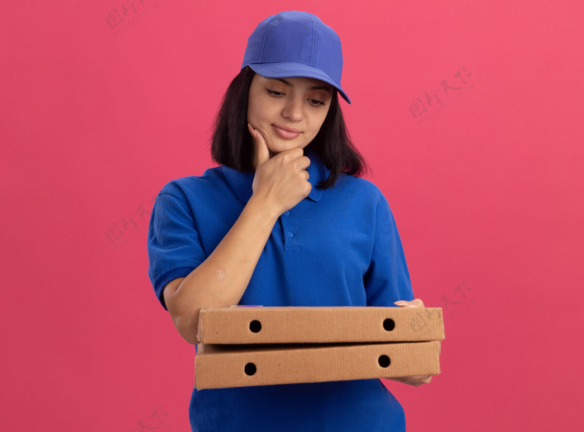 沉思身穿蓝色制服 头戴鸭舌帽 拿着披萨盒的年轻送货女孩站在粉色的墙上 神情沉思地看着披萨盒站着女孩披萨