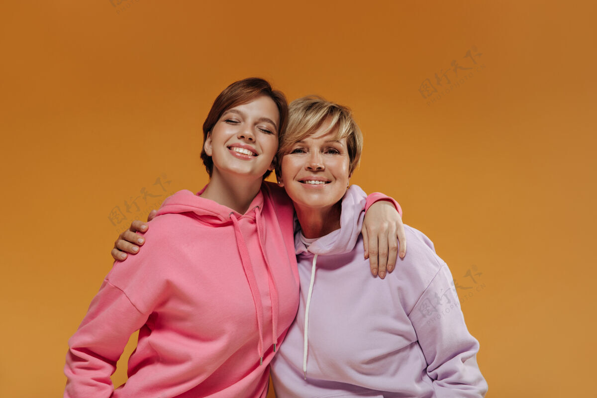 成人美丽的两位女士 现代短发 宽大明亮的连帽衫 在孤立的橙色背景下微笑着拥抱着老年人室内两个