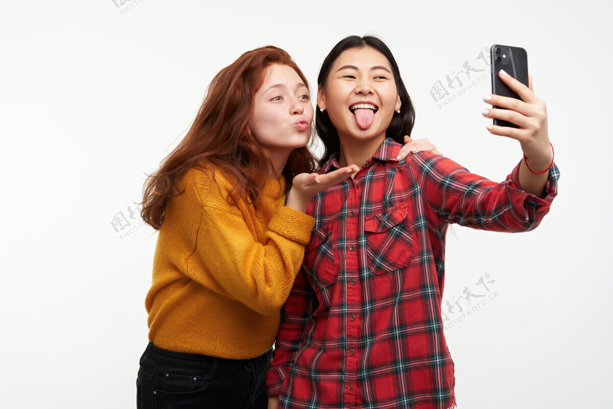 20多岁亚洲人和白种人朋友的肖像穿着黄色毛衣和格子衬衫送空气吻和展示舌头 在智能手机上自拍站在白色的墙上孤立舌头模特姜