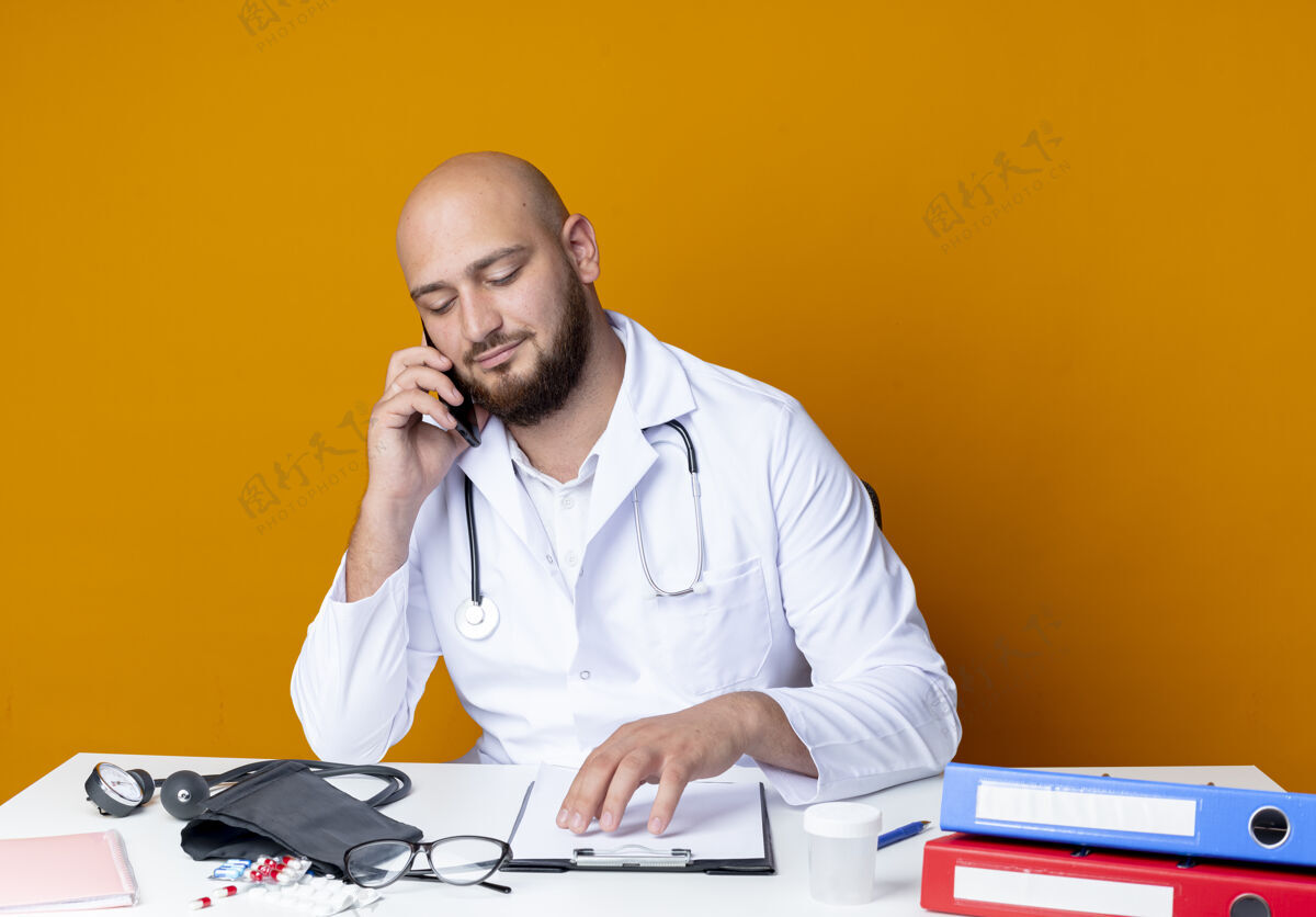 坐着高兴的年轻秃顶男医生穿着医用长袍和听诊器坐在办公桌前 手持医疗工具 在橙色背景下讲电话穿说话秃头