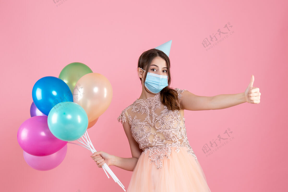 标志前视图困惑的女孩 戴着派对帽和医用面罩 举着五颜六色的气球 做着竖起大拇指的手势粉红色向上举行