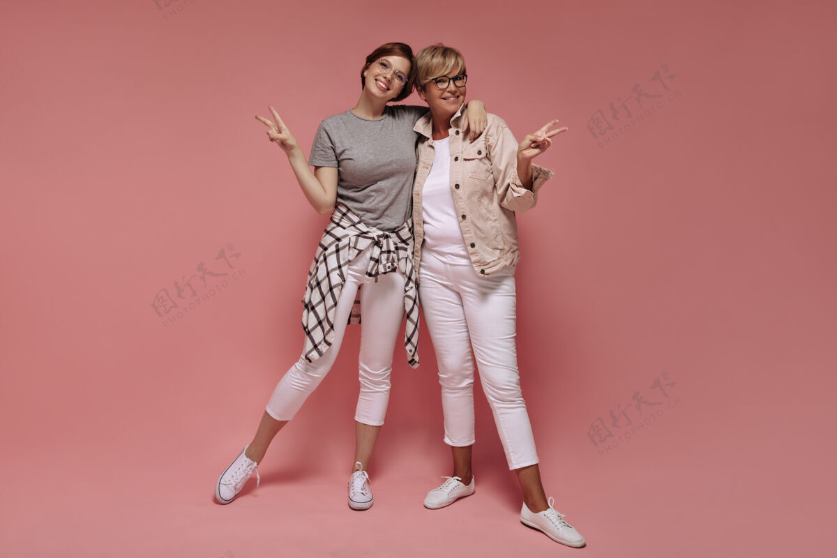 灰色两个漂亮的女人 短发 戴着现代眼镜 穿着白色紧身裤和轻便运动鞋 微笑着 在粉色的背景上展示着和平的标志白色中年衬衫