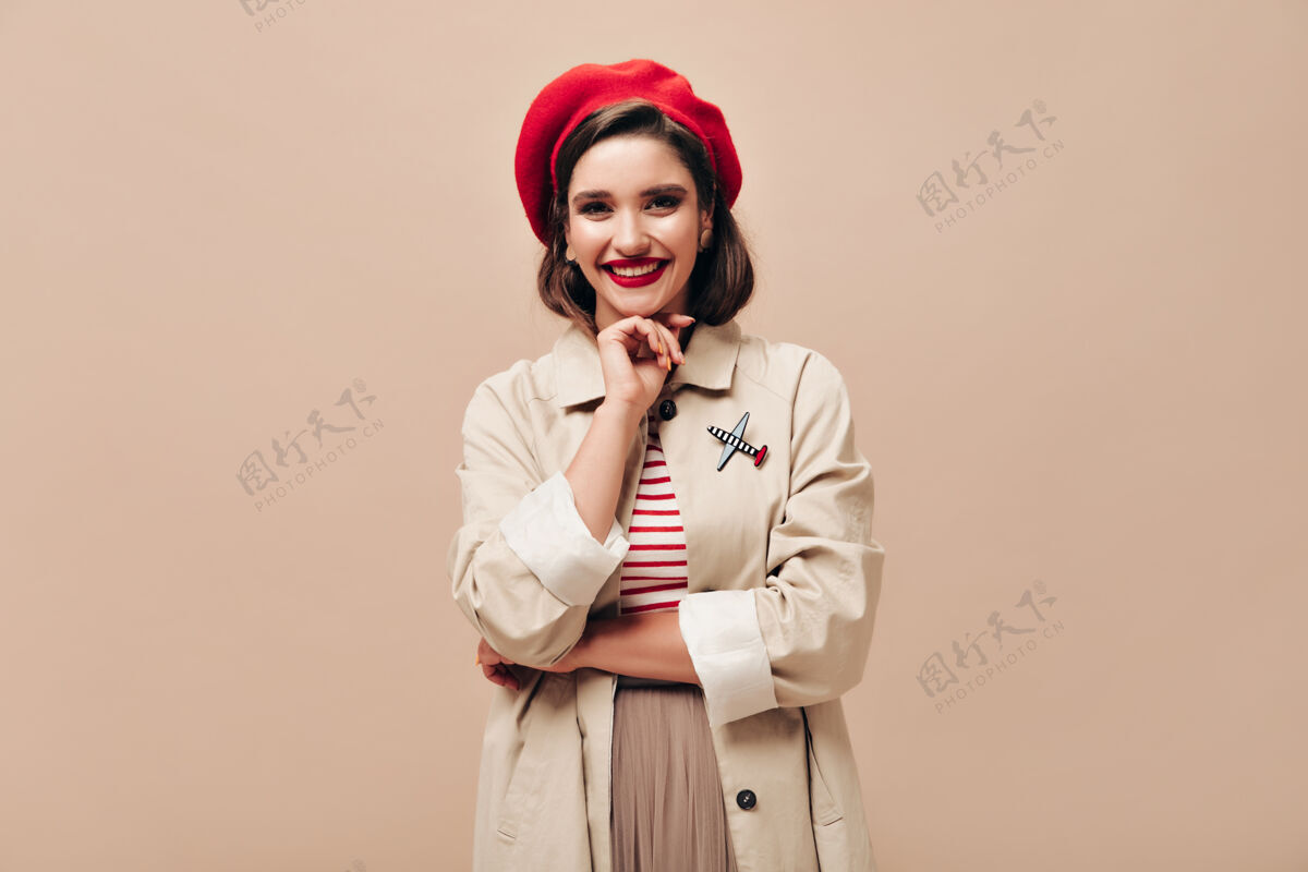 贝雷帽身着红色贝雷帽和战壕的漂亮女士在米色背景下微笑戴着红帽子 穿着轻便外套的快乐的年轻女子正对着镜头微笑站立红唇黑发