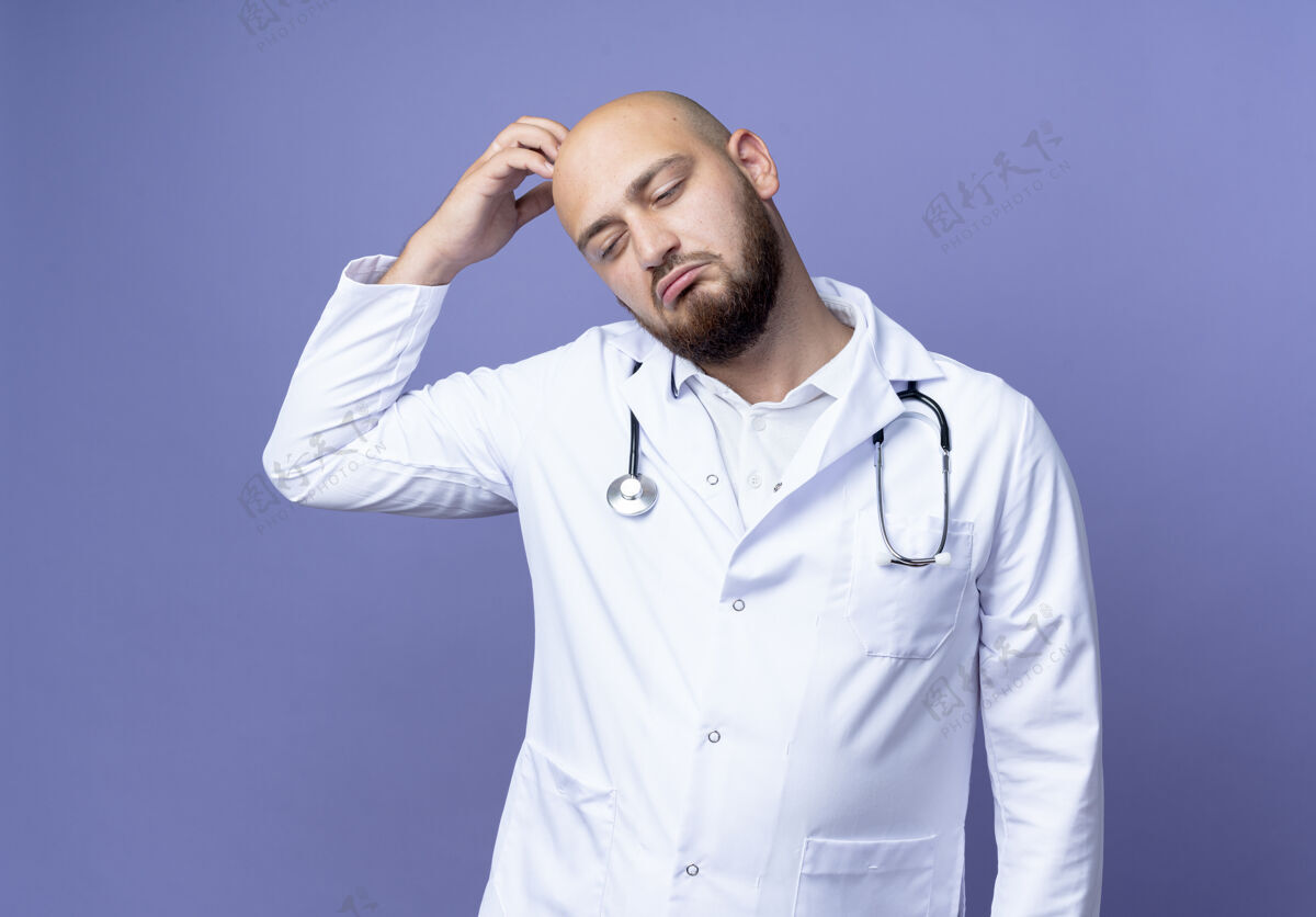医生低头看着困惑的年轻秃头男医生穿着医用长袍和听诊器在蓝色背景上搔头羽绒服男性长袍