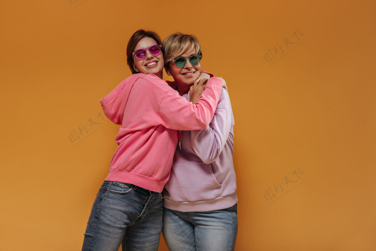 成熟现代短发女士 带着修剪的微笑和粉色和绿色的眼镜 在橙色隔离的背景下穿着宽大的时尚连帽衫牛仔裤休闲老年人