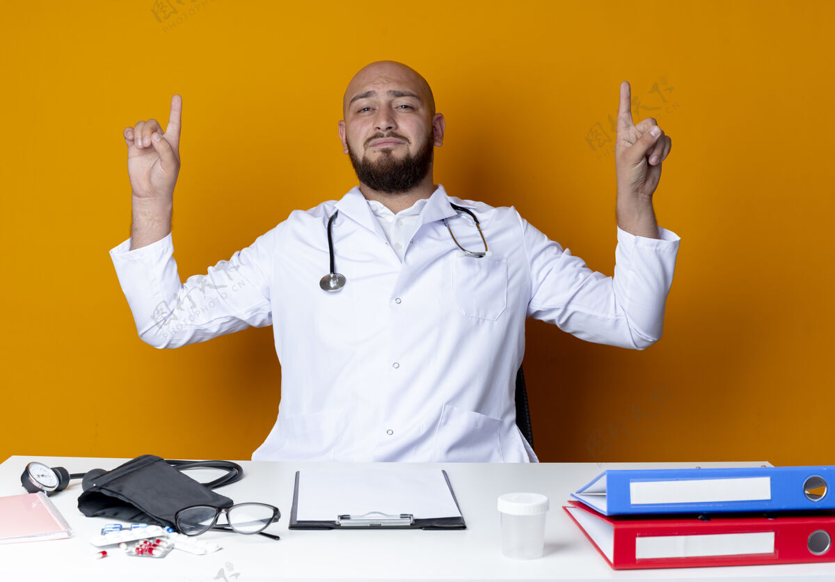 请高兴的年轻秃头男医生穿着医用长袍和听诊器坐在工作台上 用医用工具指着上面孤立的橙色背景听诊器坐着长袍