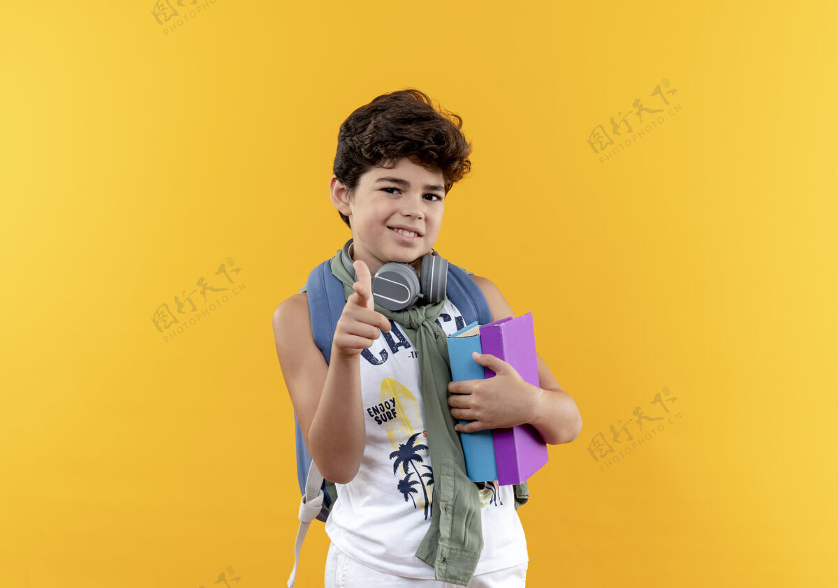 手势高兴的小男生背着书包 戴着耳机 拿着书 在黄色的背景上向你们展示孤立的手势抱小男孩