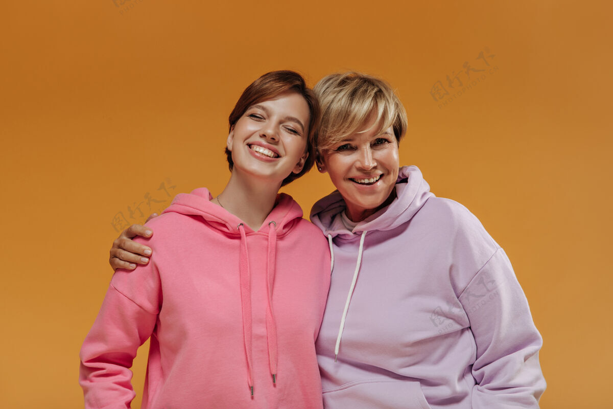 橙色背景两位时尚女性 现代短发 时尚粉色连帽衫 在孤立的橙色背景下微笑拥抱成熟粉色中等