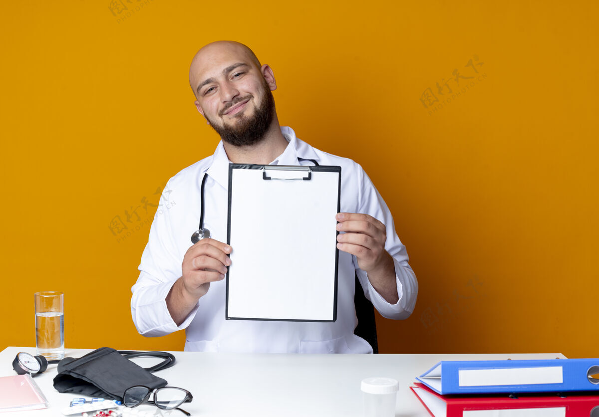 听诊器高兴的年轻秃头男医生穿着医用长袍和听诊器坐在工作台与医疗工具举行剪贴板隔离在橙色的背景工作剪贴板穿