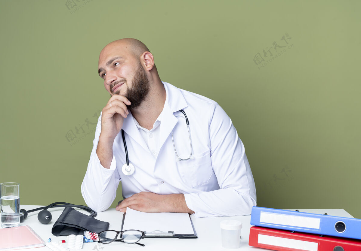 听诊器高兴的年轻秃头男医生穿着医用长袍和听诊器坐在办公桌前用医疗工具把手放在下巴上隔离在绿色的背景上长袍男性请戴
