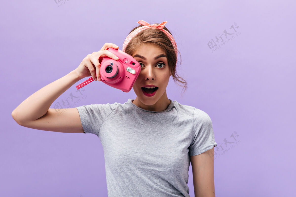女性穿灰色衣服的女孩拿着紫色背景的粉色相机戴着灰色t恤的夏日头带的年轻有趣的女人摆姿势紫色时尚肖像