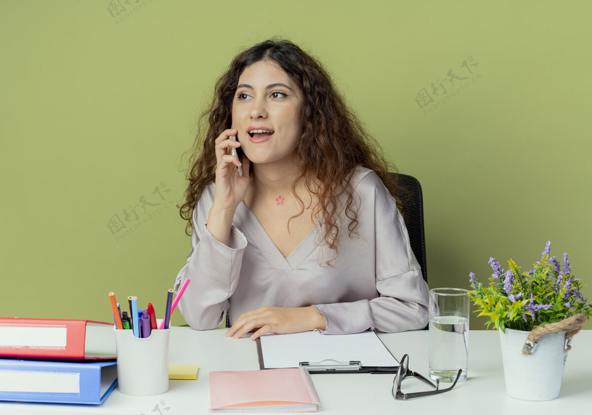 壁板看着旁边高兴的年轻漂亮的女上班族坐在办公桌旁 拿着办公工具 在橄榄色背景下讲着孤立的电话工具电话坐着
