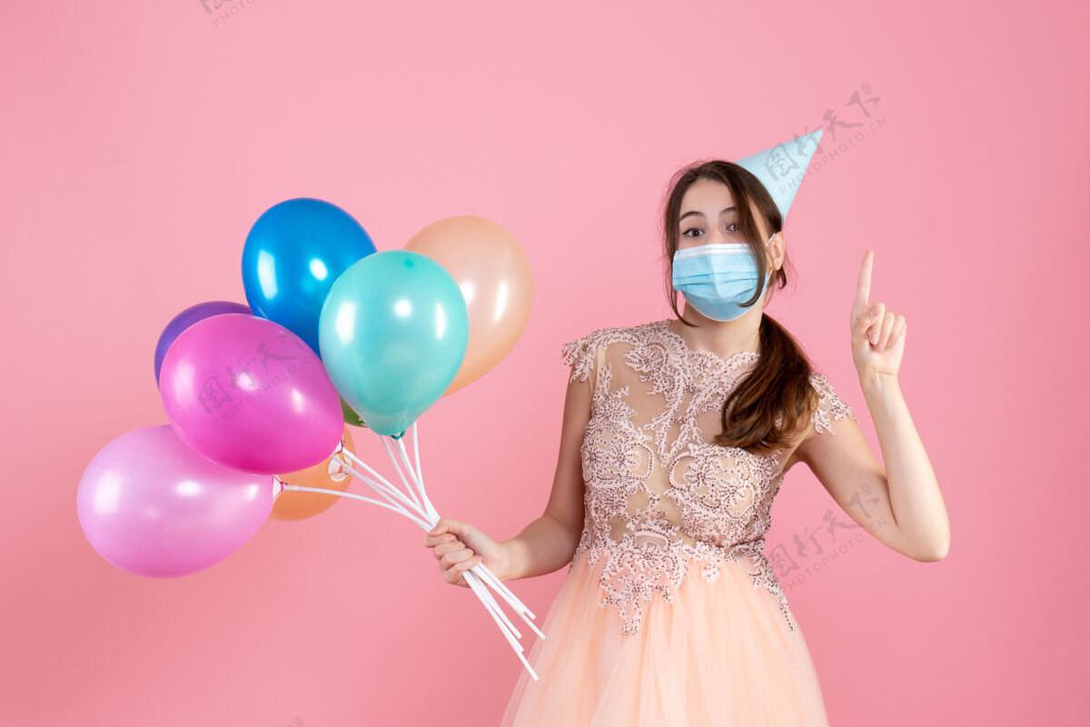 气球正面图可爱的派对女孩 戴着派对帽 拿着五颜六色的气球 手指朝上粉色快乐指向
