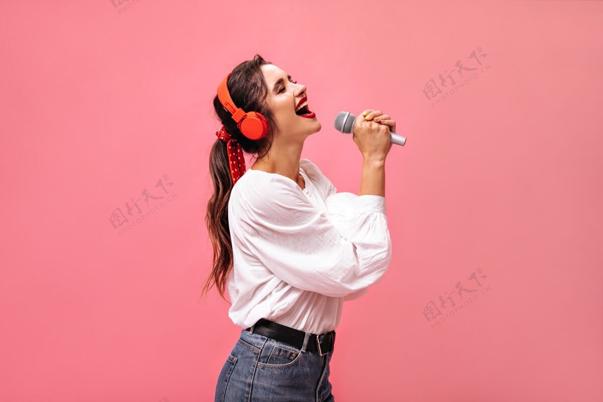 一个戴着红色耳机的情绪化女士在麦克风里唱歌穿着白色衬衫和黑色腰带牛仔裤的漂亮女孩摆姿势复制空间时尚牛仔裤