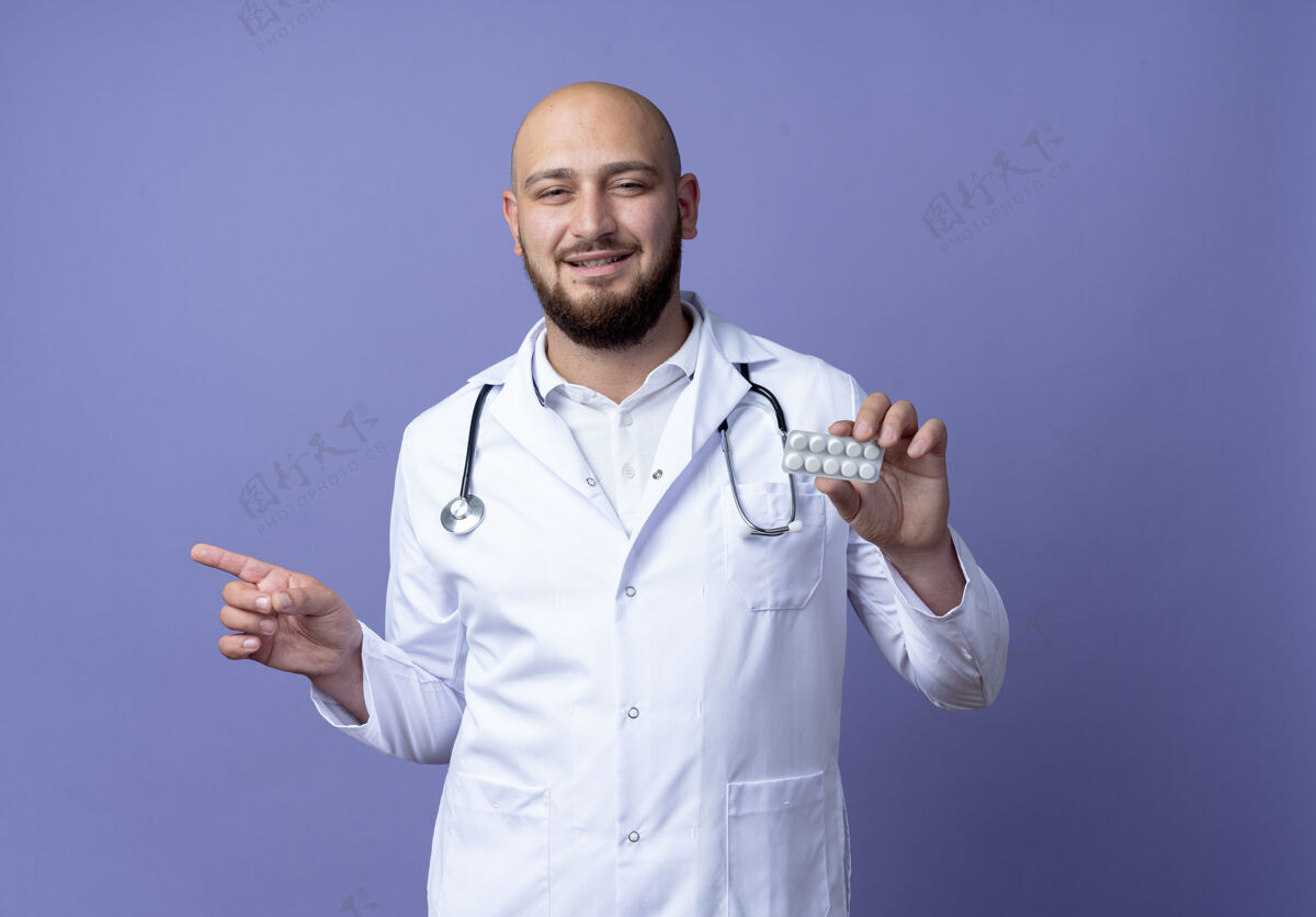 听诊器高兴的年轻秃头男医生穿着医用长袍和听诊器拿着药片和点在一边隔离在蓝色背景与复制空间医疗穿秃头