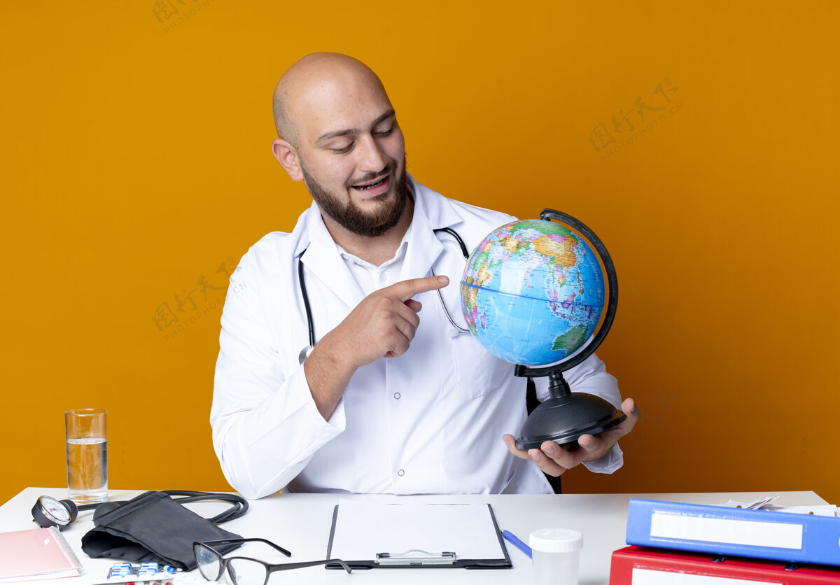 请高兴的年轻秃头男医生穿着医用长袍和听诊器坐在工作台上 手持医疗工具 指着橙色背景上孤立的地球仪听诊器秃头长袍
