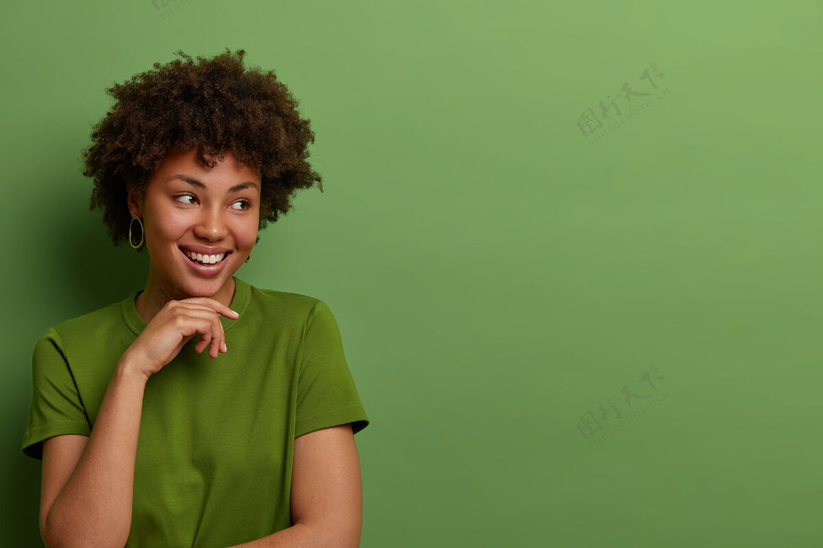外表善良乐观的美国黑人年轻女子将目光放在一边 将手轻轻放在下巴下 想象精彩的事件 穿着休闲的绿色t恤 在室内摆姿势 空白处为您的广告做准备期待享受旁白