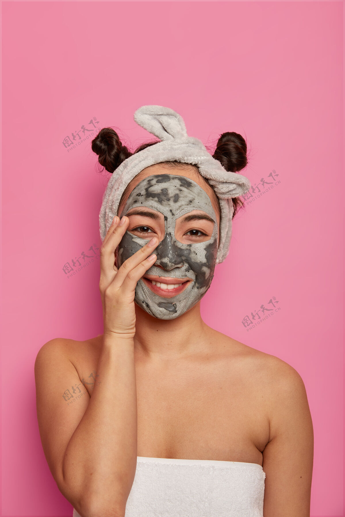 头带家里的保健和美容程序高兴的自然女人摸脸 戴上美容面膜站着 戴着头带 有两个发结 高兴地刷新皮肤 隔离在粉红色的墙上新鲜涂抹成人