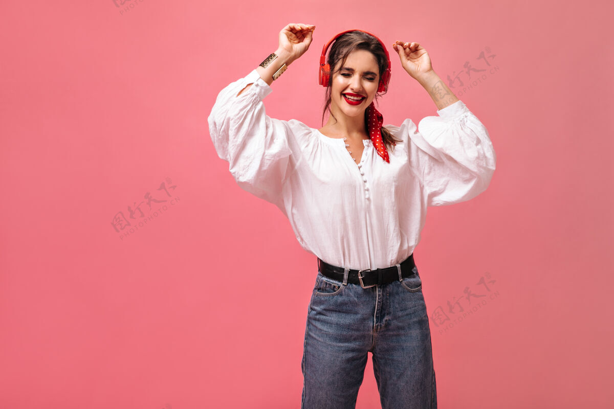 深色穿着白色上衣和牛仔裤的女士戴着耳机跳舞和听音乐时髦的女士穿着时髦的服装在粉色背景下摆姿势复制空间白色衬衫休闲