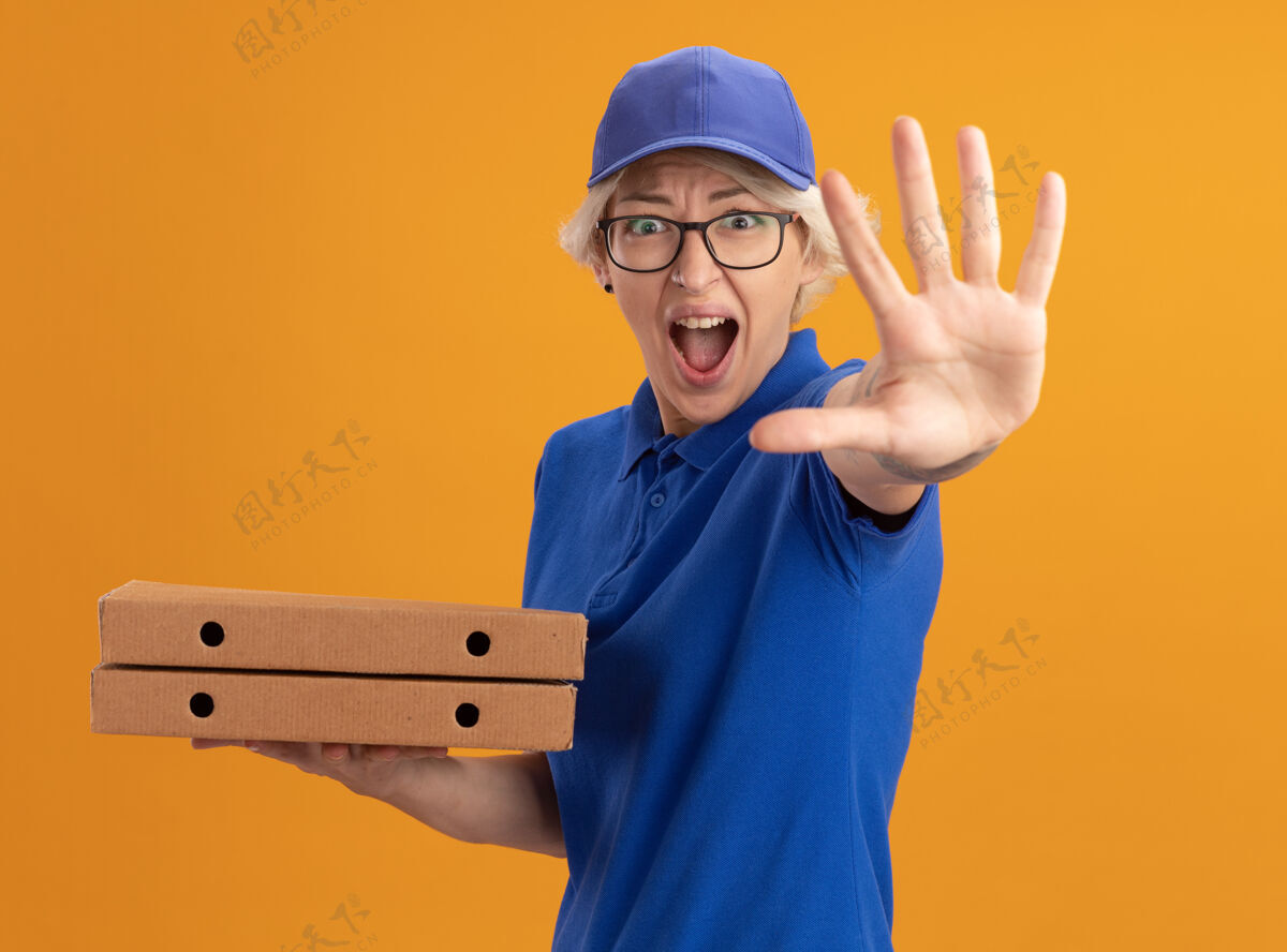 喊身穿蓝色制服 戴着眼镜 拿着比萨饼盒的年轻送货员在橙色的墙上喊着“停车”的手势制服做帽子