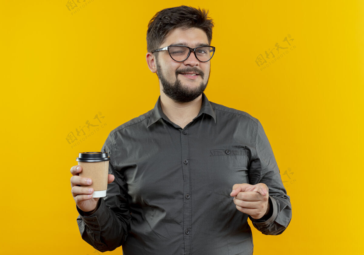 眨眼戴着眼镜的年轻商人眨着眼睛 拿着一杯咖啡 在黄色背景上孤立地向你展示手势戴着展示商人