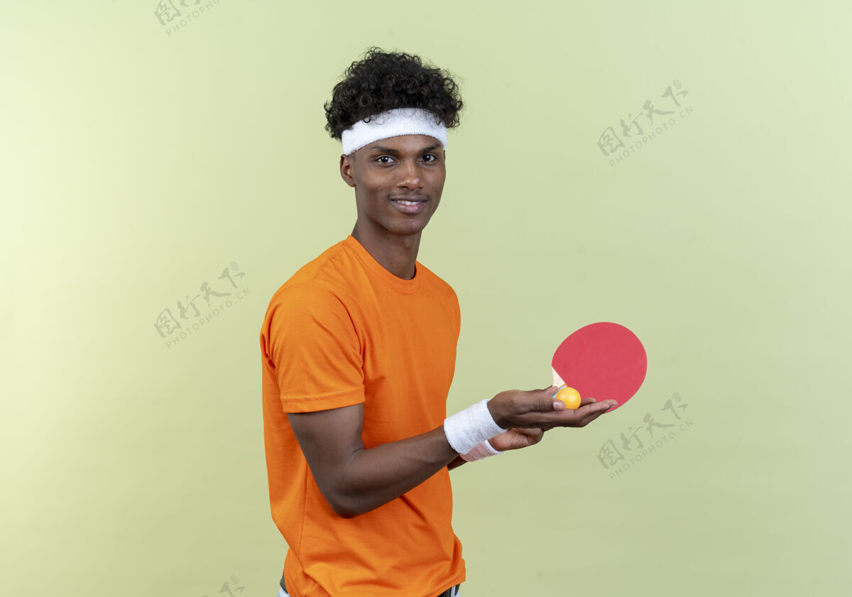 穿高兴的年轻黑人美国运动男子戴着头带和手环举行乒乓球拍与球隔离在绿色背景请腕带乒乓球