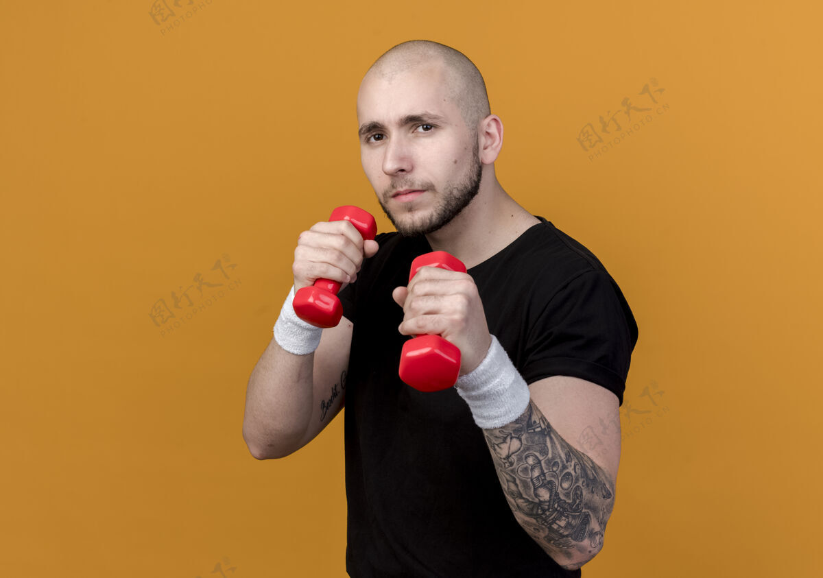 站着自信的年轻运动型男子戴着手环 举着哑铃 站在橙色背景下的格斗姿势穿着打斗姿势