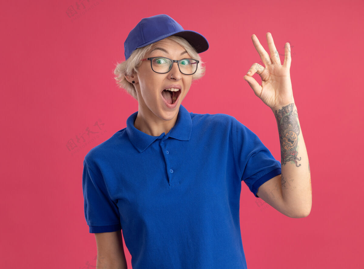 秀身着蓝色制服 头戴鸭舌帽的快乐的年轻女送货员惊讶地在粉红色的墙上展示ok标志惊喜年轻女人