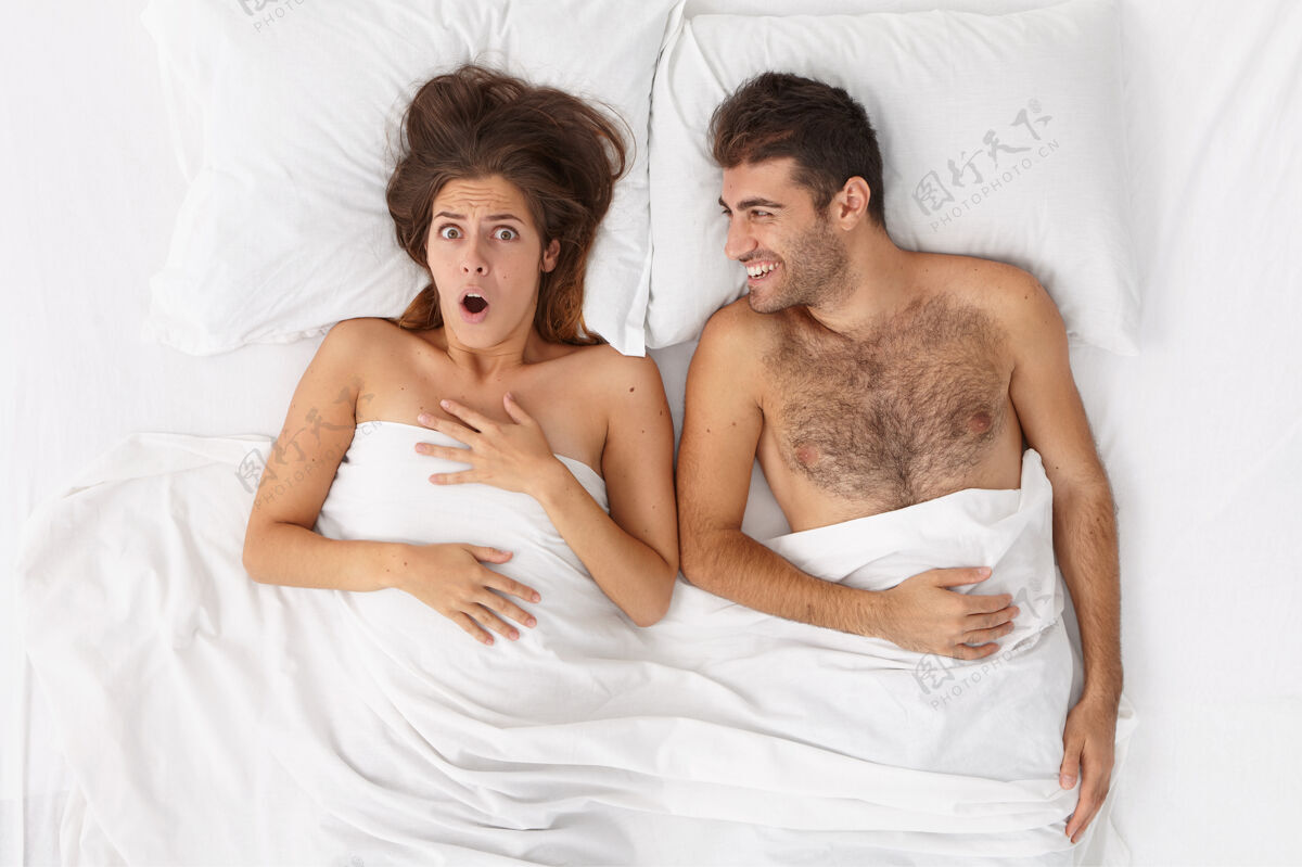 朋友年轻的新婚夫妇早上醒来受惊的女人回忆起一些惊人的事情 开朗的丈夫躺在舒适的床旁 白色床单下人 家 关系 床上用品的概念积极高兴睡眠