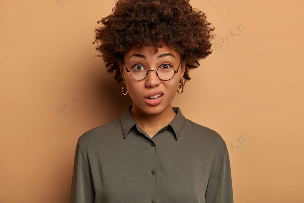 个人不满的非洲裔年轻女子表示轻蔑和怀疑 满脸不满地笑 戴着透明眼镜和衬衫 在室内对着棕色的墙壁摆姿势傻笑严重休闲