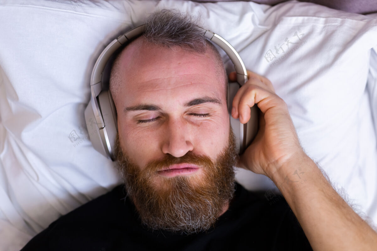舒适快乐的白人男子戴着耳机躺在床上听他最喜欢的音乐 独自休息 跳舞房子移动无线