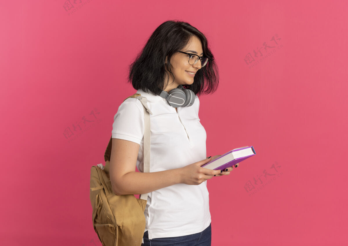 女学生年轻的高加索女孩 面带微笑 戴着眼镜 戴着耳机 背着书包 站在一旁 手里拿着书和笔记本 粉色的有复印空间空间拿着年轻