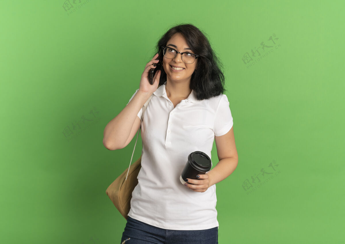 杯子年轻的高加索女孩 面带微笑 戴着眼镜 背着书包 拿着咖啡杯在绿色的草地上讲着电话漂亮复制电话