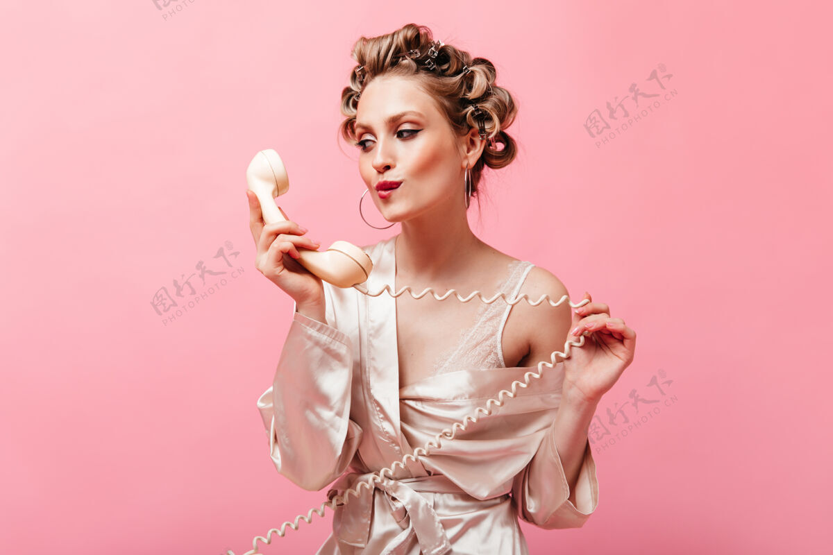 长袍穿着丝绸家装的漂亮女人看着手机 在粉红色的墙上摆姿势金发享受家庭主妇