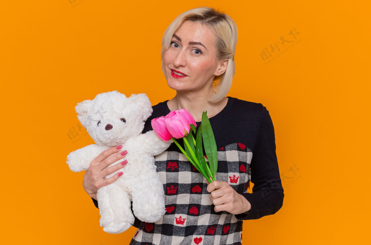 郁金香身着漂亮连衣裙的快乐年轻女子手持一束郁金香和泰迪熊作为礼物 站在橙色的墙上微笑着看着前方庆祝国际妇女节熊礼物站