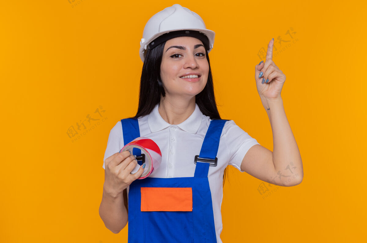 年轻人身着建筑制服 戴着安全帽 拿着胶带的年轻建筑工人自信地微笑着 食指站在橙色的墙上信心展示站着