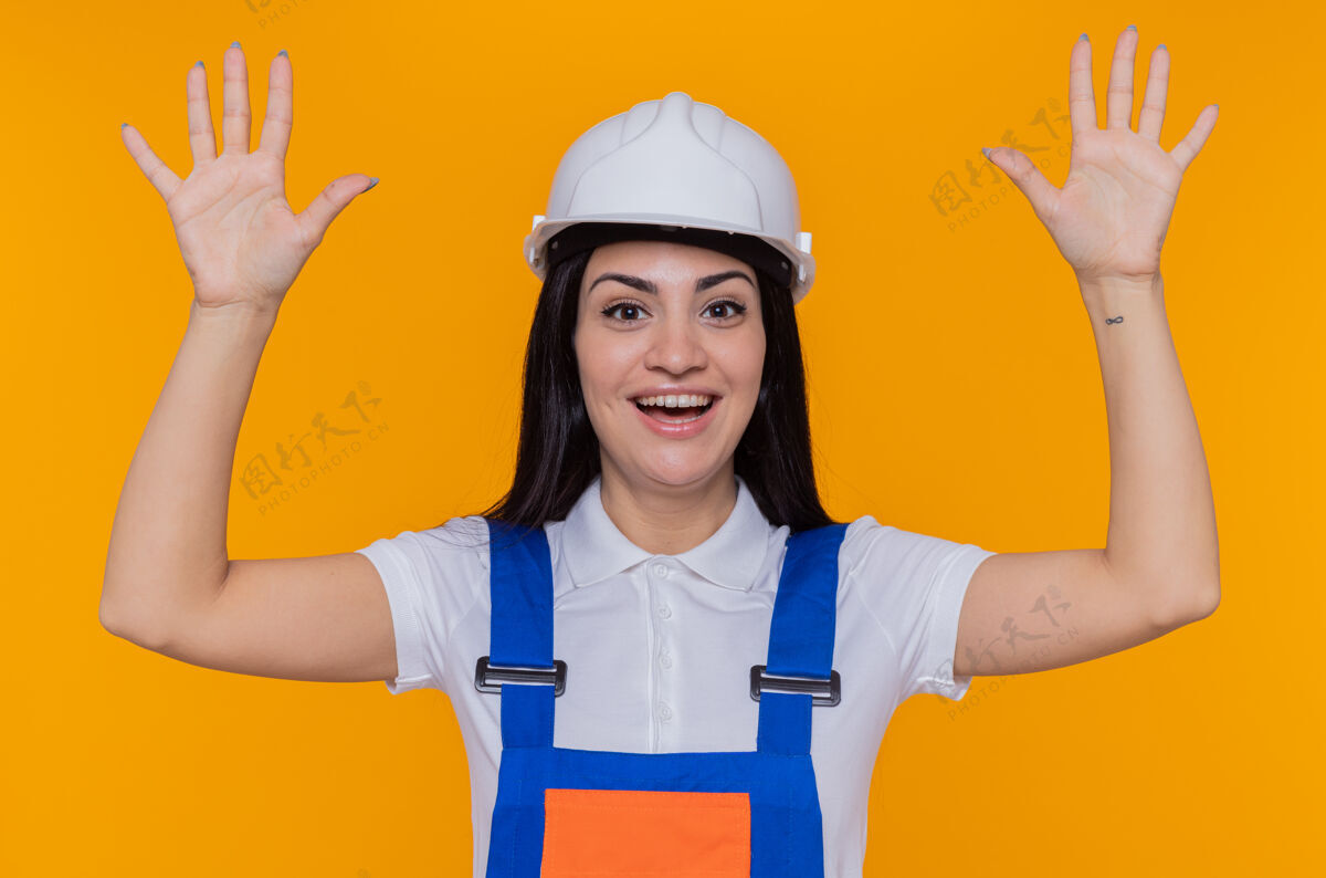 建设者身穿施工制服 头戴安全帽的年轻建筑女工人站在橙色的墙上 面带微笑 开心 积极地站在前面举起年轻快乐