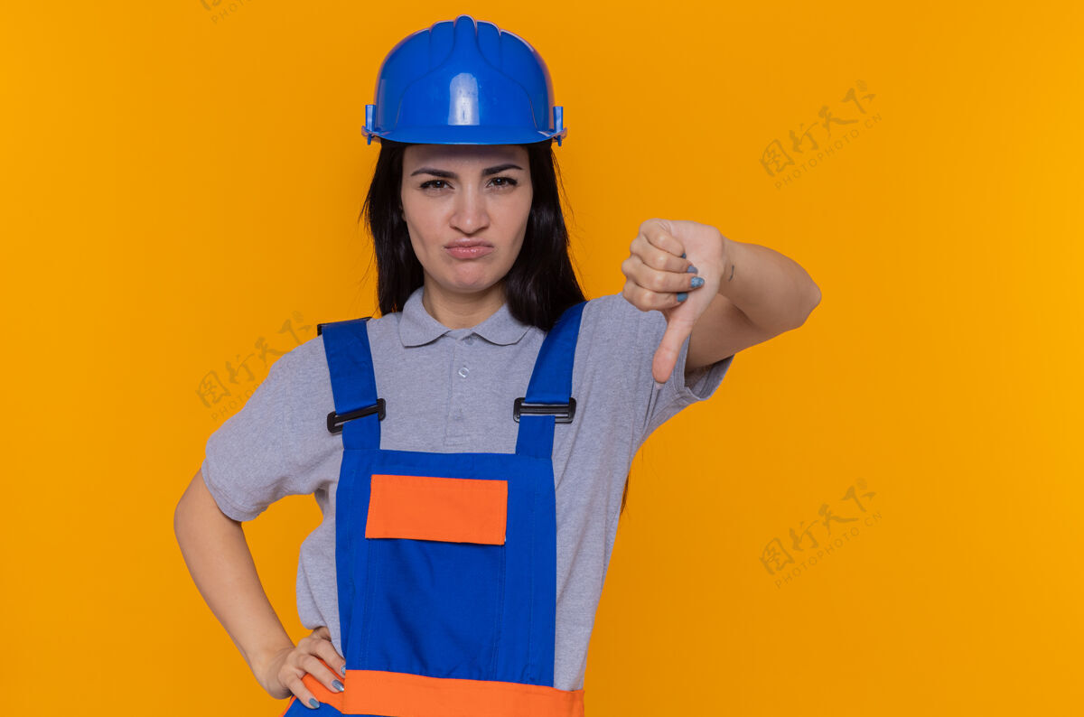 拇指身穿建筑制服 头戴安全帽的年轻建筑工人不高兴地朝前看 站在橙色的墙上竖起大拇指不高兴制服站着
