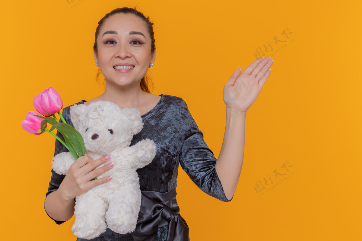 三月快乐的亚洲女人手持一束粉红色的郁金香和泰迪熊 站在橙色的墙上欢笑着庆祝国际妇女节亚洲欢呼泰迪