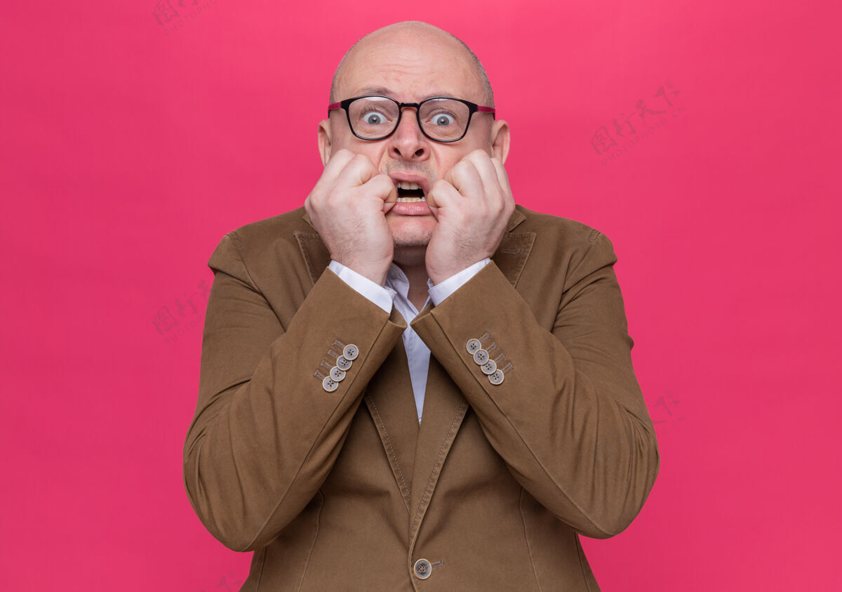 秃头穿着西装的中年秃头男人戴着眼镜 看着站在粉色墙壁上紧张紧张 咬牙切齿的指甲咬男人紧张