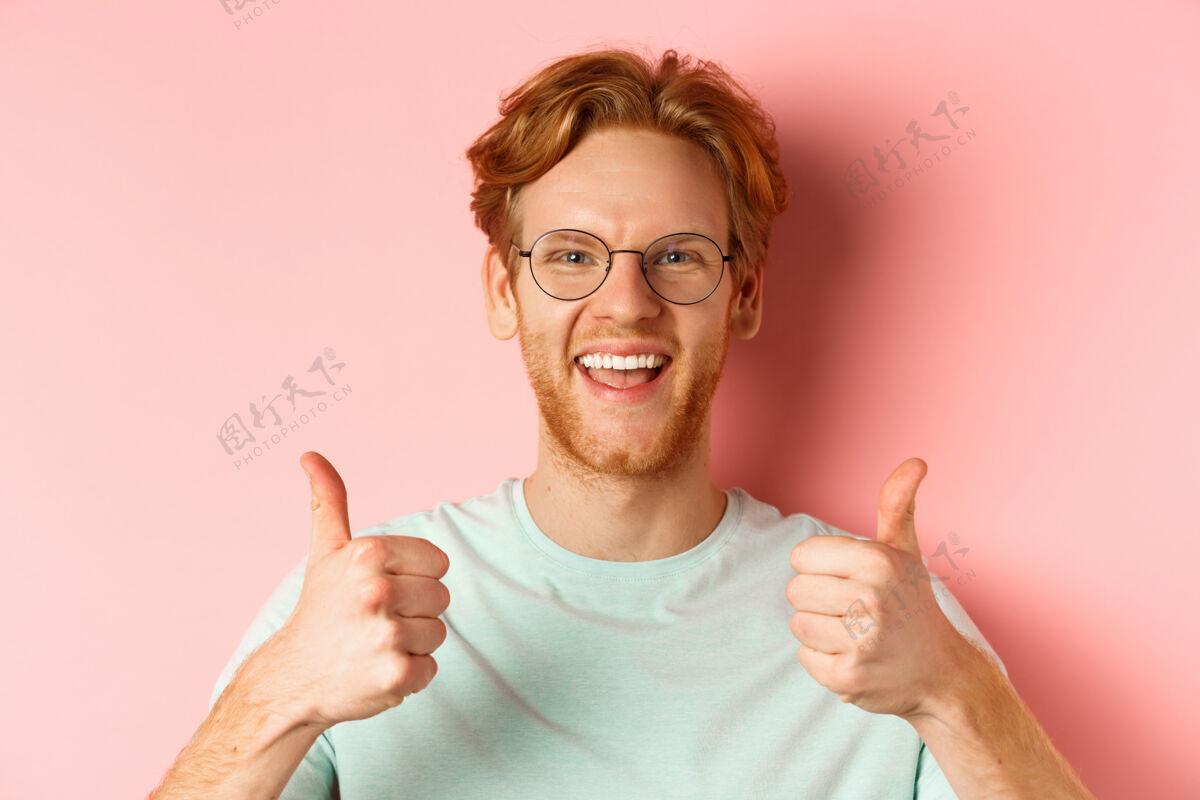 红发满意的男顾客脸上竖起大拇指表示赞同 微笑着高兴 戴着眼镜和t恤 粉色背景喜欢崇拜学生