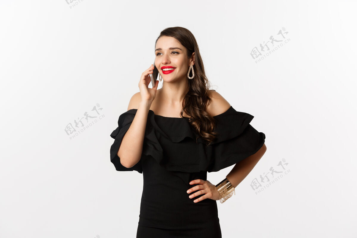 女人成功的年轻女性穿着黑色礼服 红色唇膏和化妆品 在手机上微笑着 站在白色背景上女人 时尚 美丽 莫比尔 优雅 服装 模特 口红 女性 年轻 美丽 表情 魅力 女性 电话 时尚 黑发