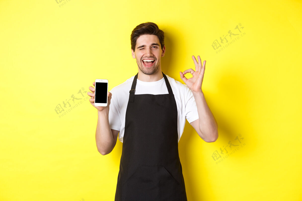 男士帅气的咖啡店员工展示ok标志和智能手机屏幕 推荐应用程序 站在黄色背景上模特自信手机