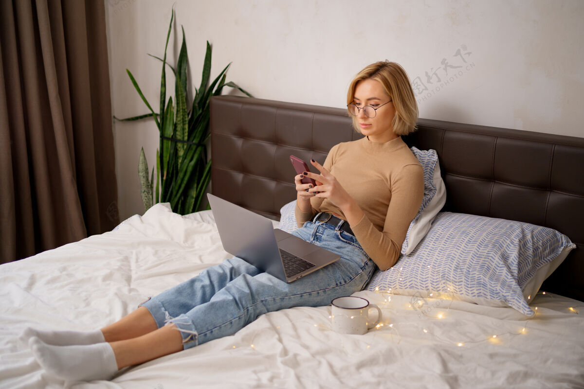 女人一个金发短发的女人穿着牛仔裤躺在白色的床上 手提电脑里拿着智能手机牛仔裤卧室金发